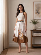 A woman models a sleeveless two-piece outfit featuring a cream crop top with dark brown trim and a matching pleated midi skirt with a brown and cream color-blocked design and floral print. She is standing in a warm, neutral-toned room with a side table and curtains.