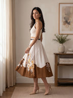 A woman models a sleeveless midi skirt with a cream bodice and a brown lower panel featuring a floral print, paired with a black belt. She is standing in a bright, modern room with a wooden side table and neutral curtains.