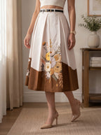 A woman models a high-waisted midi skirt with a color-blocked design. The top half is off-white with a large floral print in yellow, orange, and white, while the bottom half is solid brown.