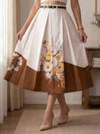 A woman models a pleated midi skirt with a color-blocked design of off-white on top and brown on the bottom, featuring a large yellow and white floral print on the front and one side panel. She is wearing beige high heels in a bright indoor setting.