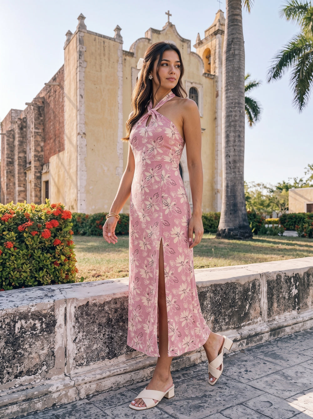 A woman wearing a pink floral halter maxi dress with a thigh-high slit, standing outdoors in front of a historic stone church and palm trees.
