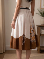 A woman wearing a pleated midi skirt with a color-blocked design. The top portion is cream with a thin black waistband, the middle is solid brown, and the lower portion features a floral print in yellow, orange, and green tones over a brown base.