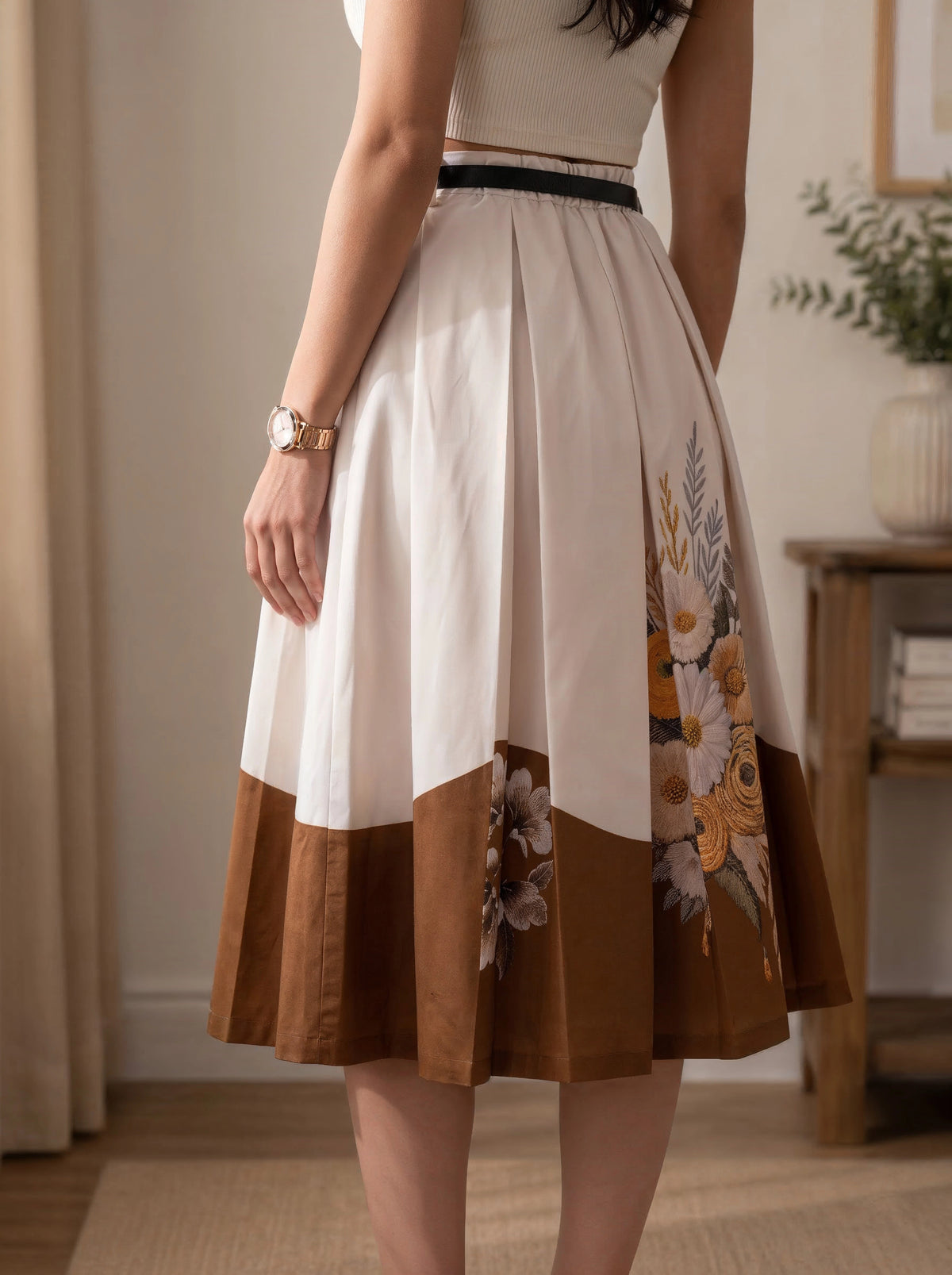 A woman wearing a pleated midi skirt with a color-blocked design. The top portion is cream with a thin black waistband, the middle is solid brown, and the lower portion features a floral print in yellow, orange, and green tones over a brown base.