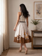 A woman stands with her back to the camera in a bright room, wearing a sleeveless midi dress with a cream bodice, a thin black waist belt, and a brown skirt featuring a floral print and a solid brown border.