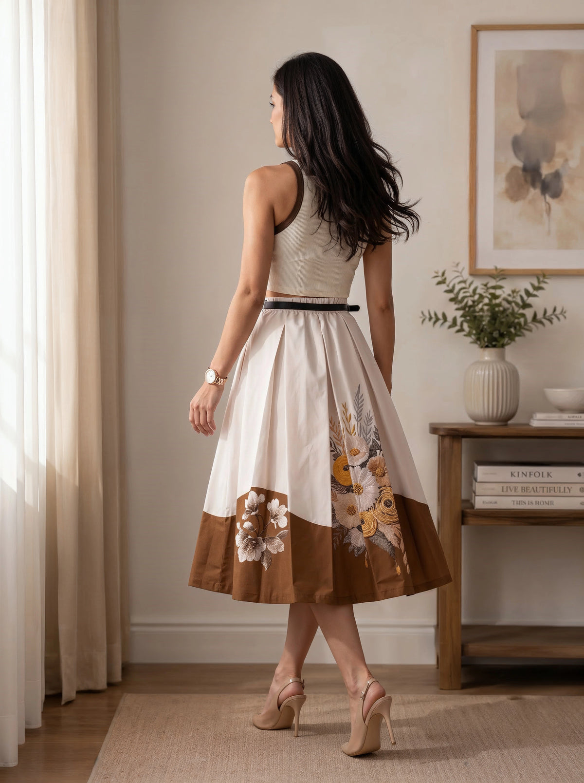 A woman stands with her back to the camera in a bright room, wearing a sleeveless midi dress with a cream bodice, a thin black waist belt, and a brown skirt featuring a floral print and a solid brown border.