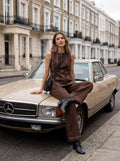 A woman models a brown sleeveless top and matching wide-leg trousers with black ankle boots, sitting on the hood of a beige Mercedes-Benz car on a city street.