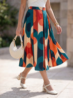 A woman walking outdoors wearing a high-waisted midi skirt with a bold abstract geometric pattern in teal, orange, cream, and navy. She is also wearing a white top, white Mary Jane heels, and carrying a white crescent-shaped handbag.