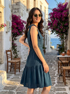 A woman wearing a sleeveless teal blue tiered dress and sunglasses stands on a cobblestone street in a Mediterranean-style town, with white buildings, purple bougainvillea flowers, and the blue sea in the background.