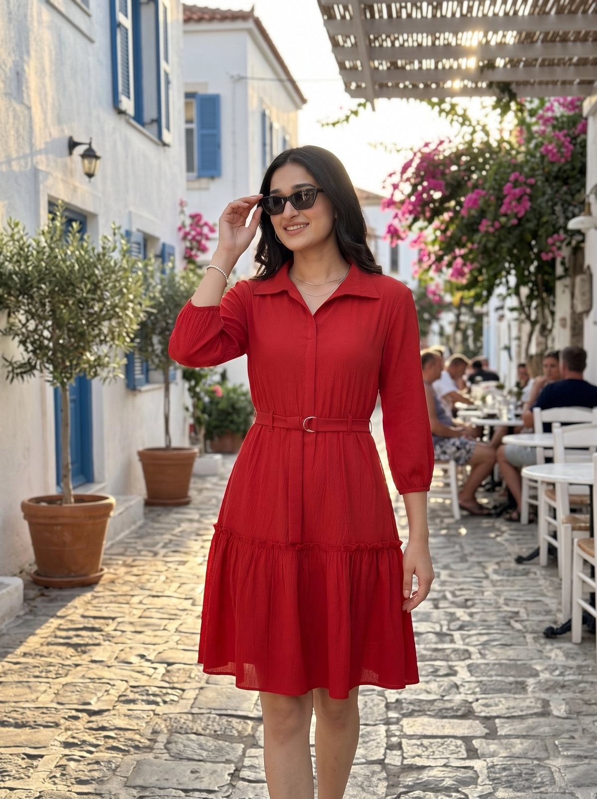 A woman wearing a bright red collared midi dress with three-quarter sleeves and a matching belt walks down a sunlit cobblestone street lined with white buildings, blue shutters, potted plants, and hanging pink flowers.