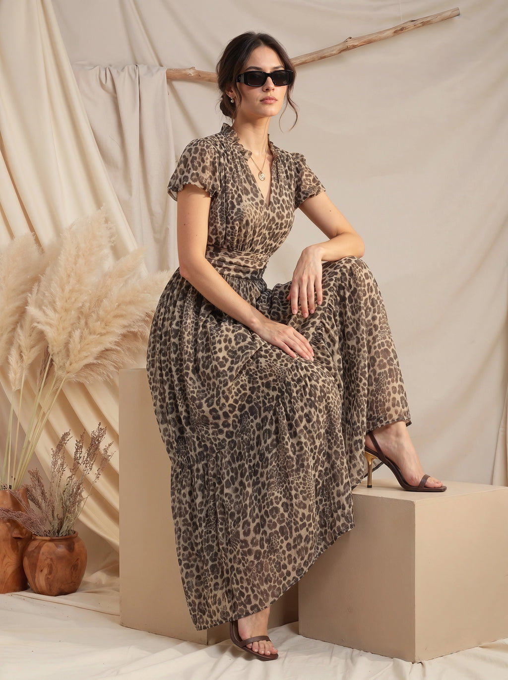 A woman wearing a leopard-print maxi dress and black sunglasses sits on a beige block against a draped cream fabric backdrop with pampas grass decor.