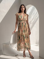 A woman models a sleeveless midi dress with a beige, brown, and green botanical print, standing in a minimalist white room with a modern white chair and strong architectural shadows.
