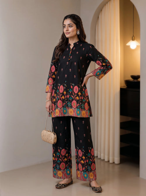 A woman models a black three-piece kurta set with vibrant multicolor floral patterns, holding a small woven handbag in a modern indoor setting.