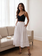 A woman modeling a black spaghetti-strap crop top and a high-waisted white midi skirt with side pockets, standing in a bright living room next to a beige sofa.