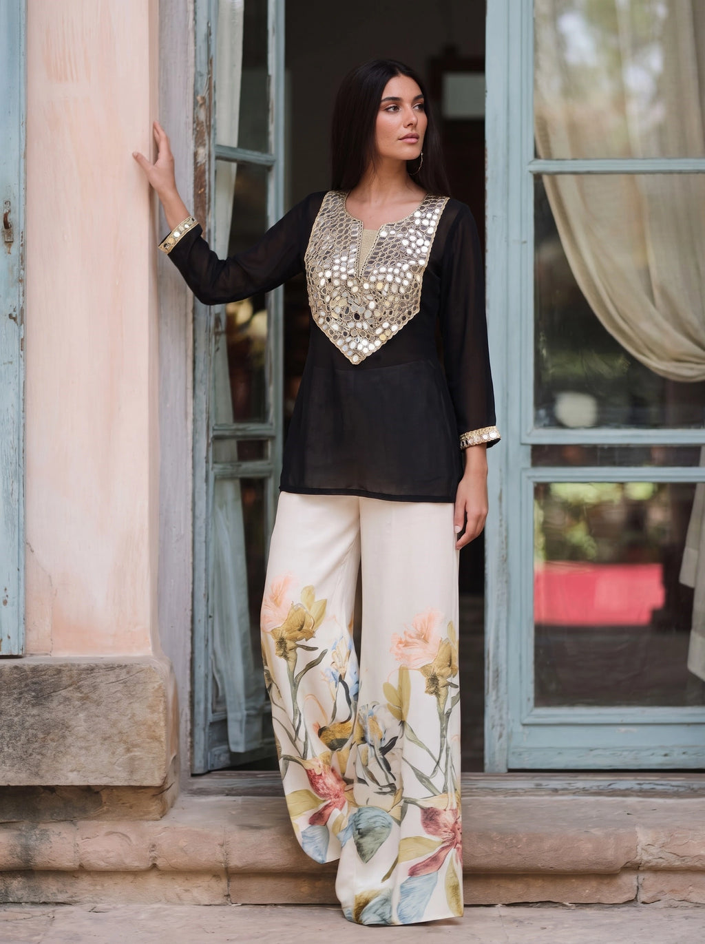 A woman models a black three-piece traditional outfit featuring a long-sleeve top with a gold sequined V-neckline, paired with wide-leg cream trousers adorned with a pastel floral print.