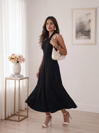 A woman modeling a navy blue tiered mini dress with elbow-length sleeves, paired with white block-heel sandals, standing in a bright room with a wooden floor and a potted plant.