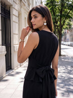 A woman wearing a sleeveless black dress with a tied waist bow, photographed outdoors on a city sidewalk with buildings and trees in the background.