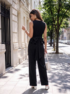 A woman stands on a city sidewalk wearing a black sleeveless wide-leg jumpsuit with a large bow detail at the back waist, holding a small white handbag.