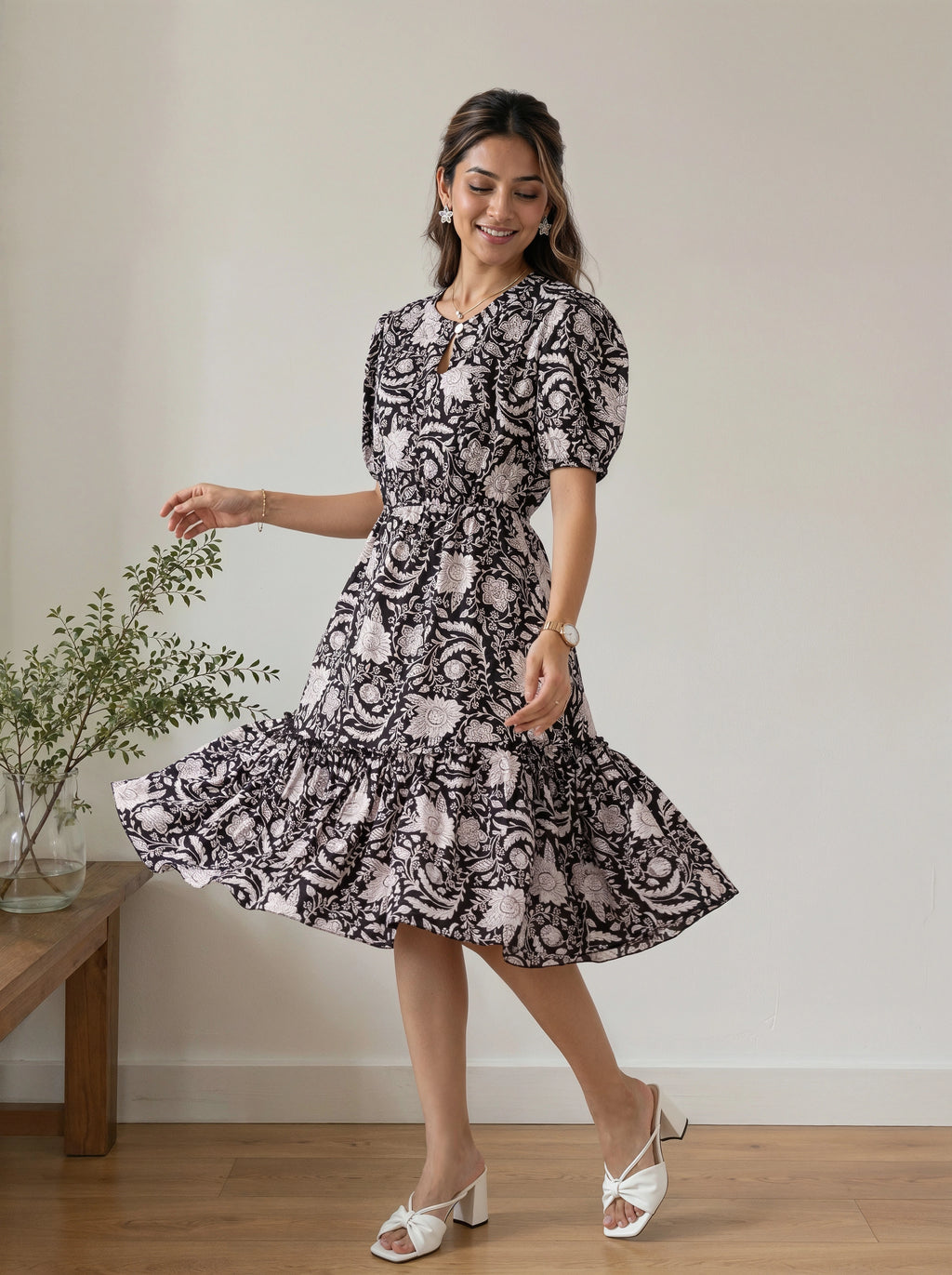 A woman modeling a short puff-sleeve midi dress with a black and white floral paisley pattern, standing in a room with a wooden floor and a small plant on a side table.