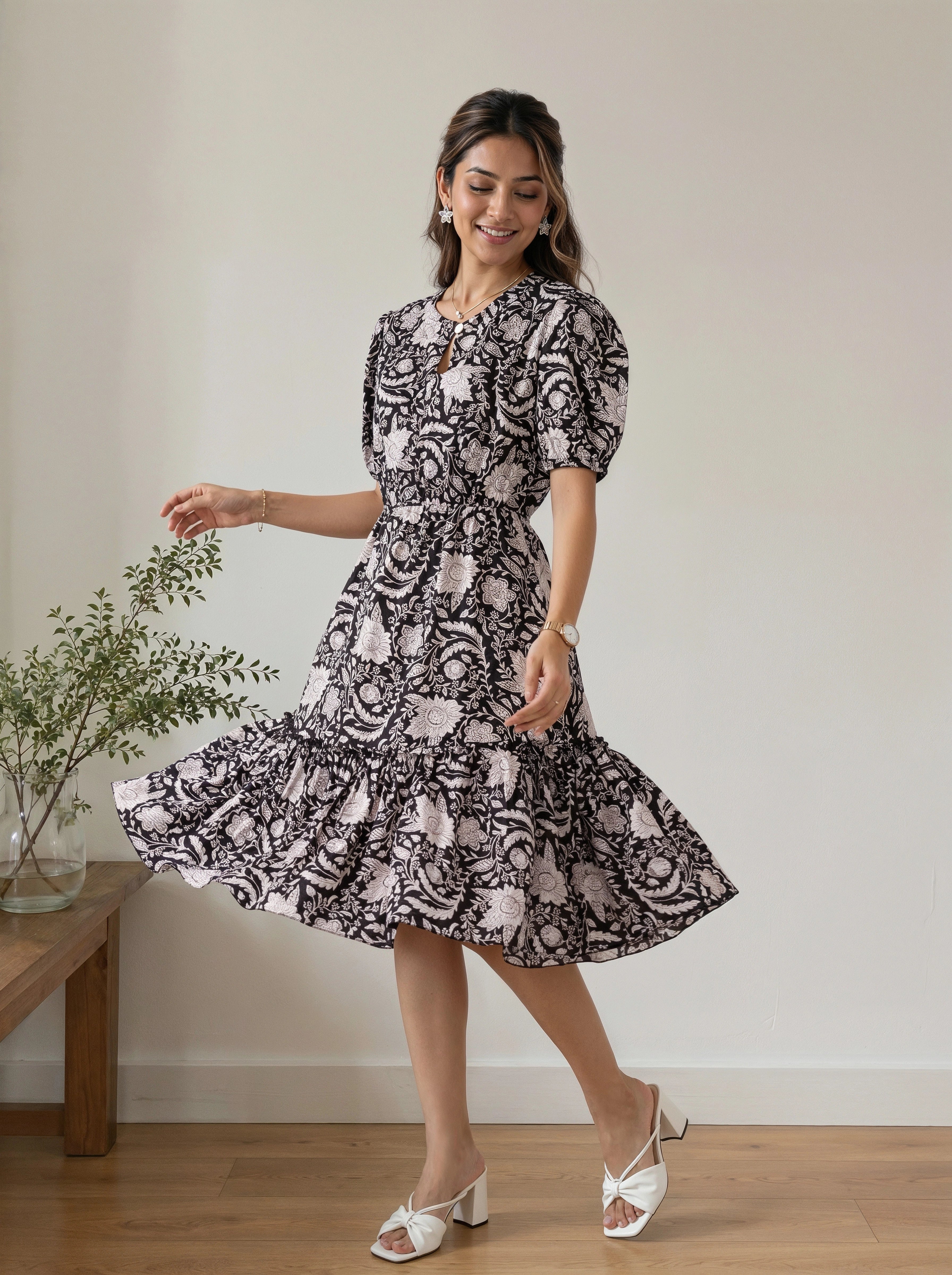 A woman modeling a short puff-sleeve midi dress with a black and white floral paisley pattern, standing in a room with a wooden floor and a small plant on a side table.