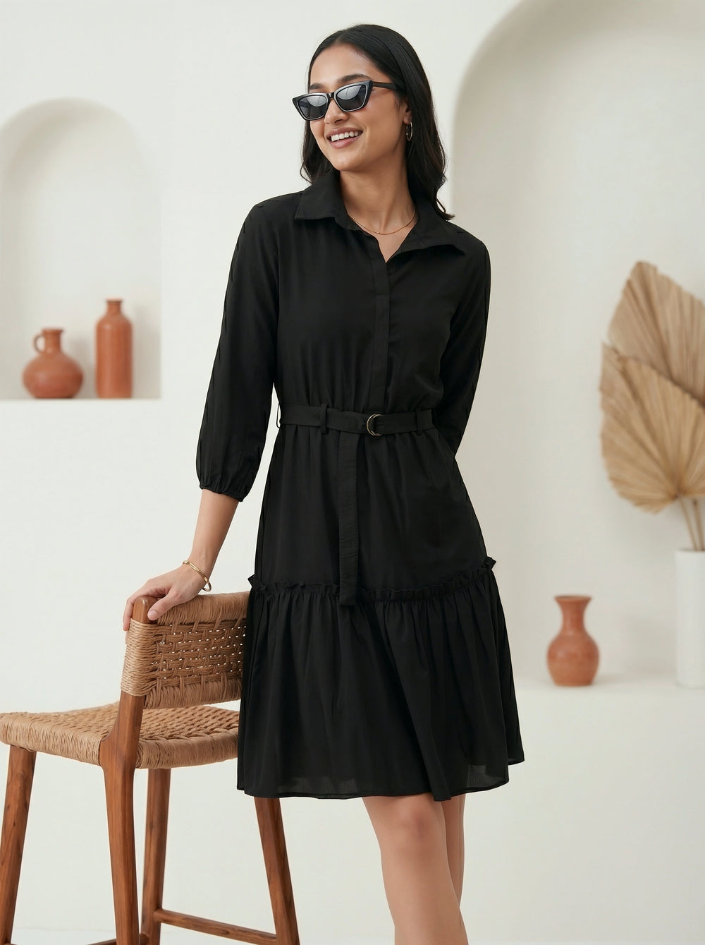 Woman wearing a black dress standing in a room with a white wall and wooden chair.