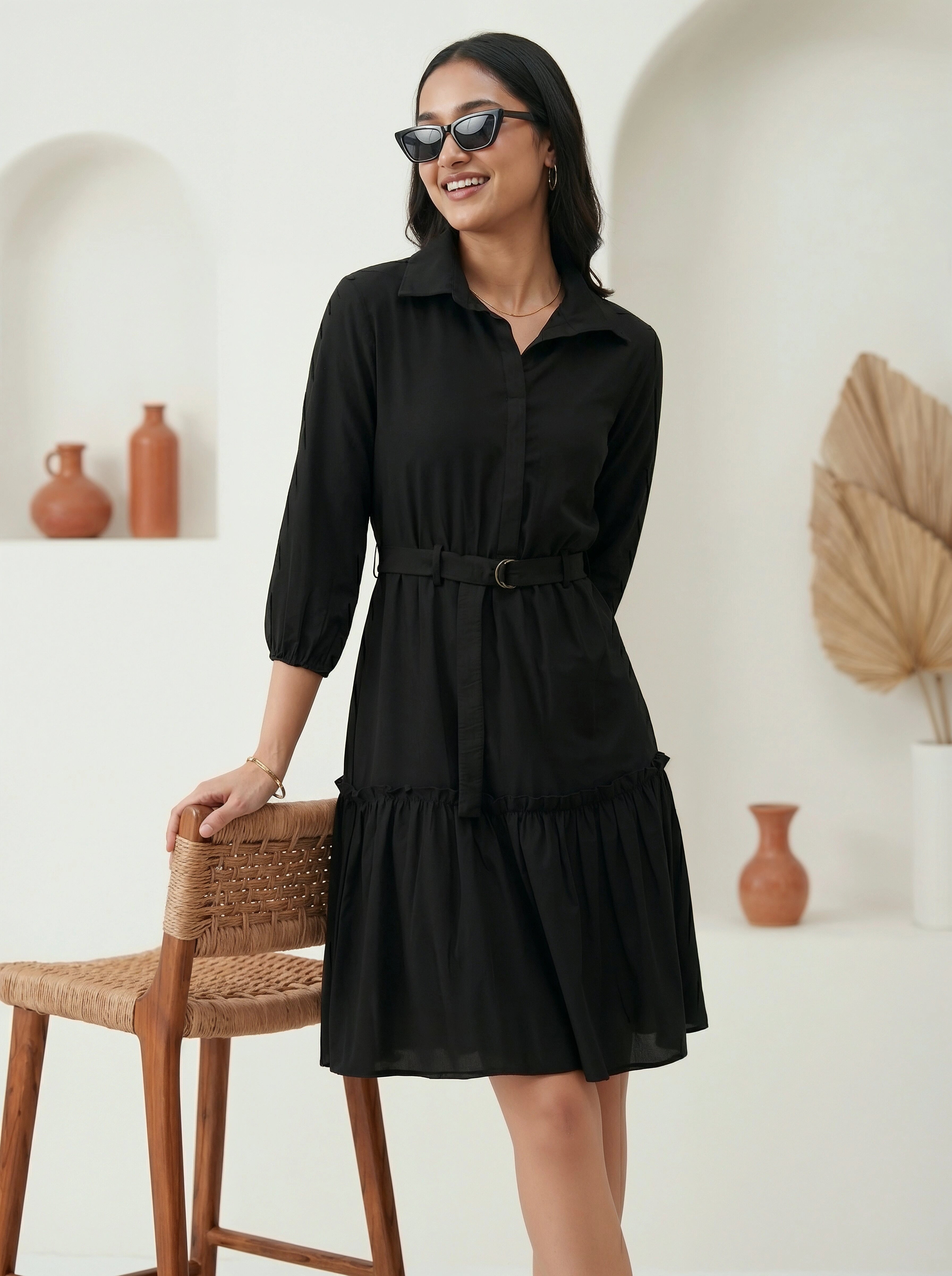 Woman wearing a black dress standing in a room with a white wall and wooden chair.