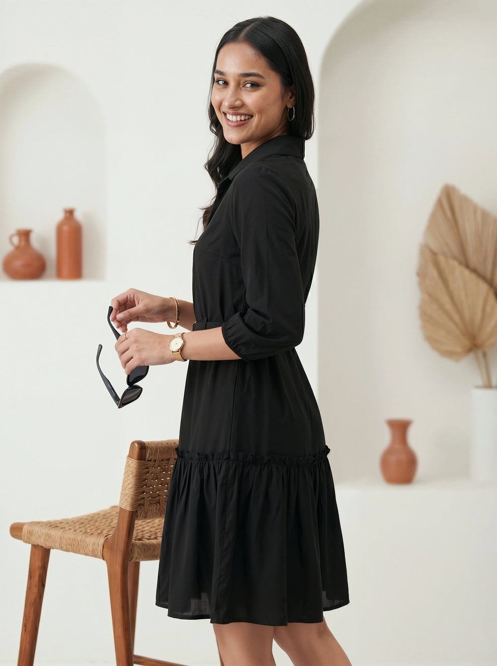 Woman in a black dress holding sunglasses in a minimalistic room.