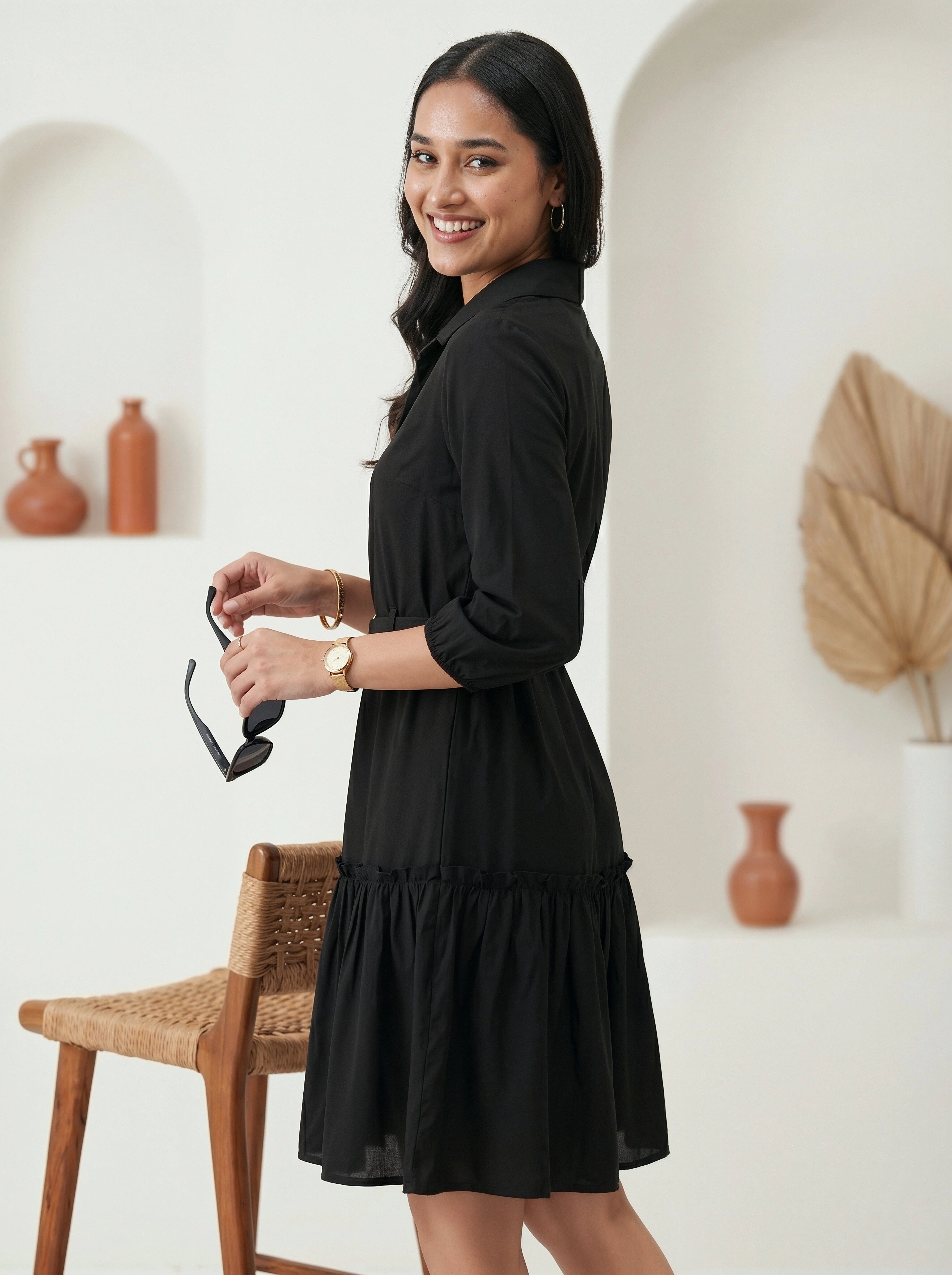 Woman in a black dress holding sunglasses in a minimalistic room.