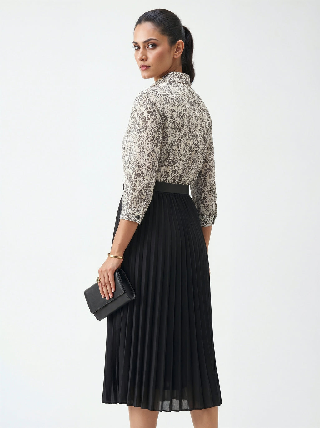 Woman wearing a patterned blouse and black pleated skirt holding a clutch on a white background