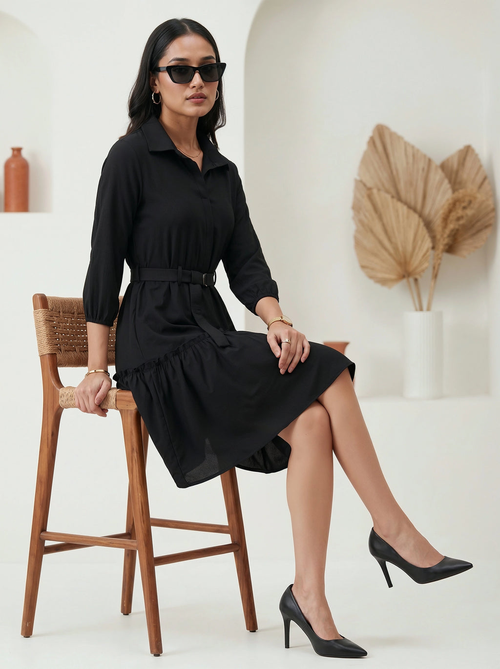 Woman in a black dress sitting on a wooden stool in a minimalistic setting.