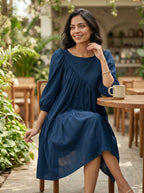 Woman in a blue dress sitting at an outdoor cafe table with plants and tables in the background.