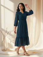 Woman wearing a blue dress standing in a softly lit room with curtains.