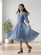 Woman in a blue dress standing in a bright room with a plant in the background