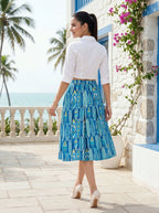 Woman in a white blouse and blue patterned skirt standing on a sunlit patio with ocean view.