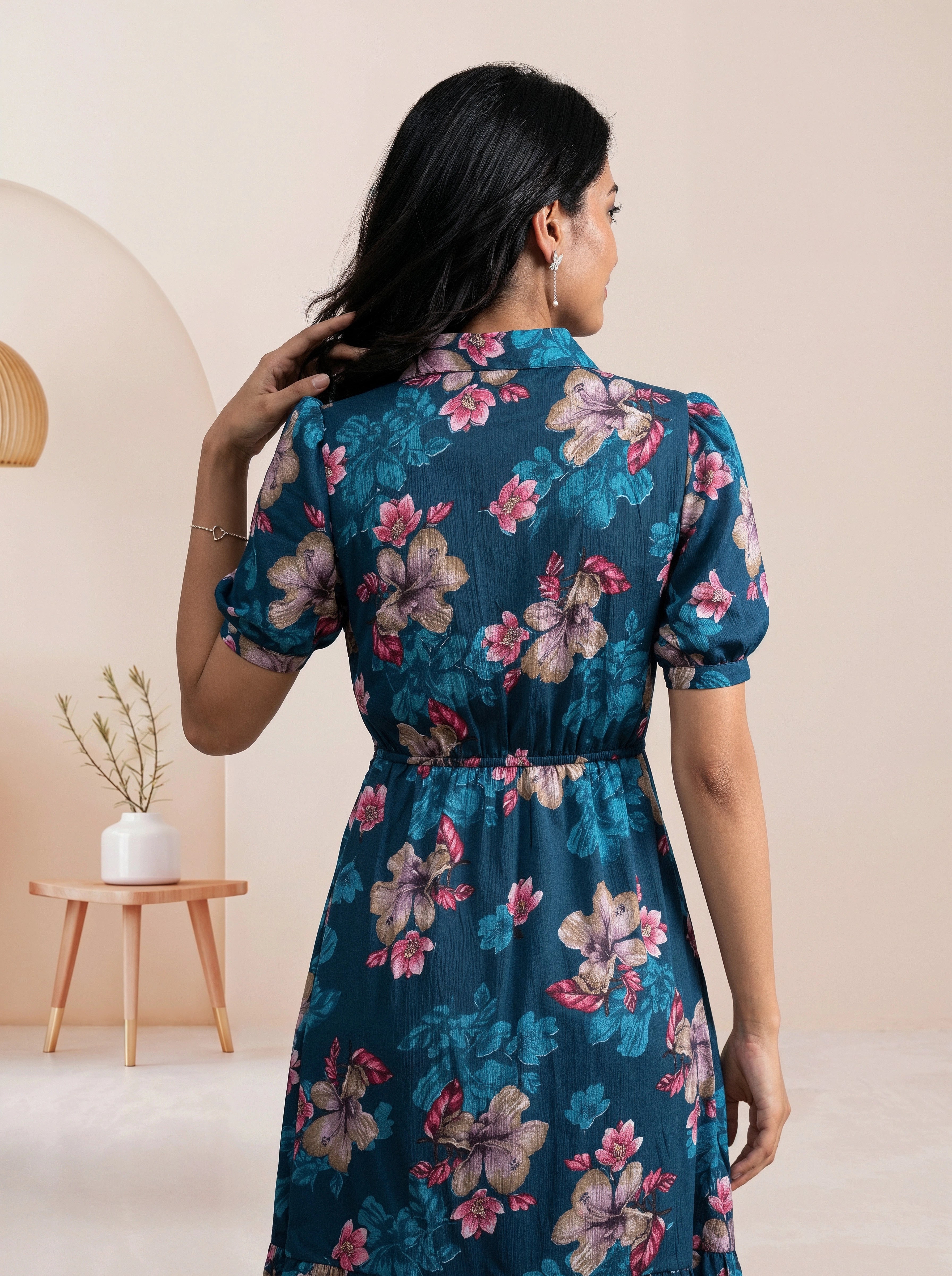 A woman wearing a short-sleeve floral dress, shown from the back in a studio setting with a beige background and a small wooden side table.