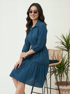 Woman wearing a blue dress standing in a room with a plant and chair.