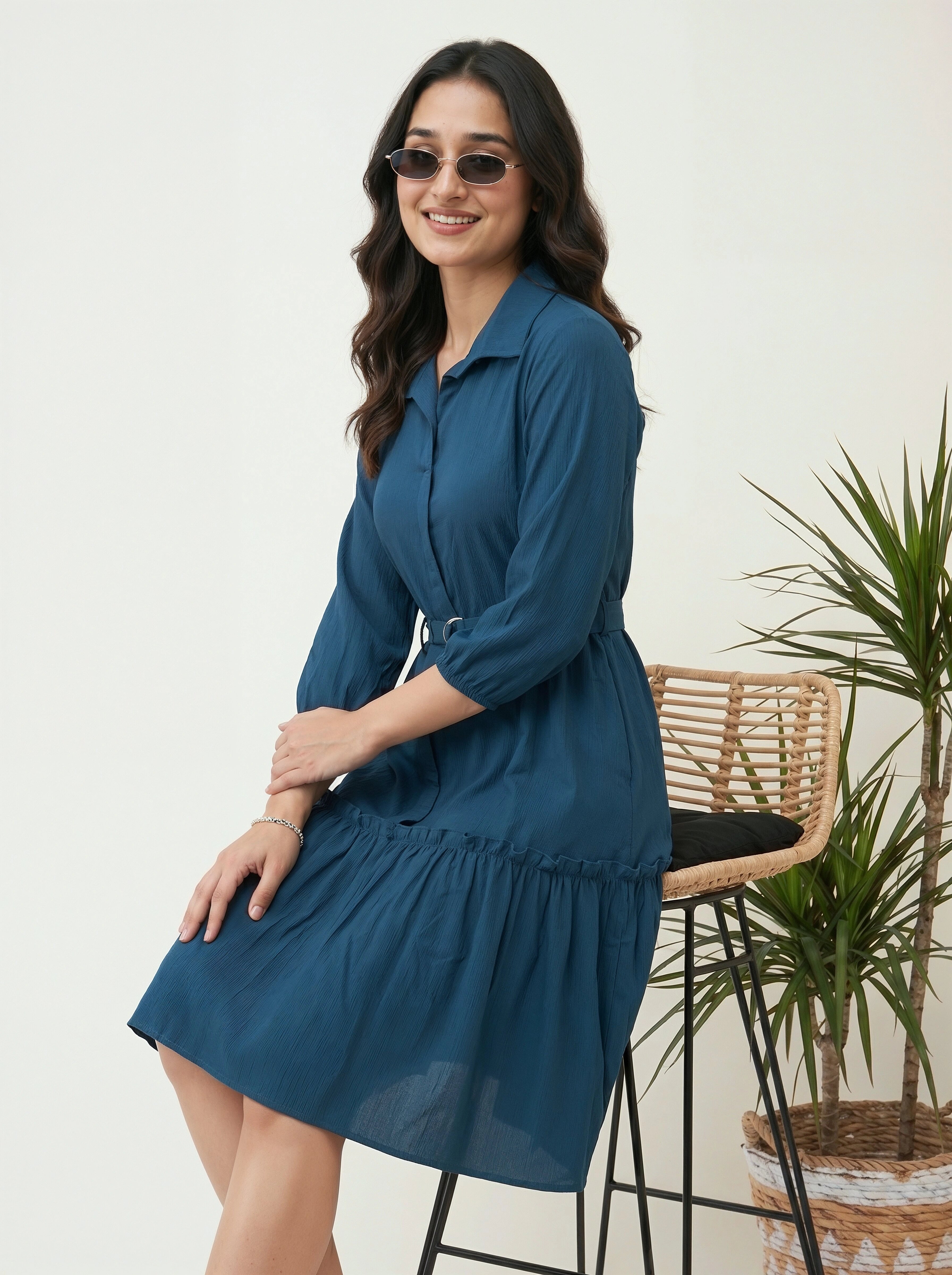 Woman wearing a blue dress standing in a room with a plant and chair.