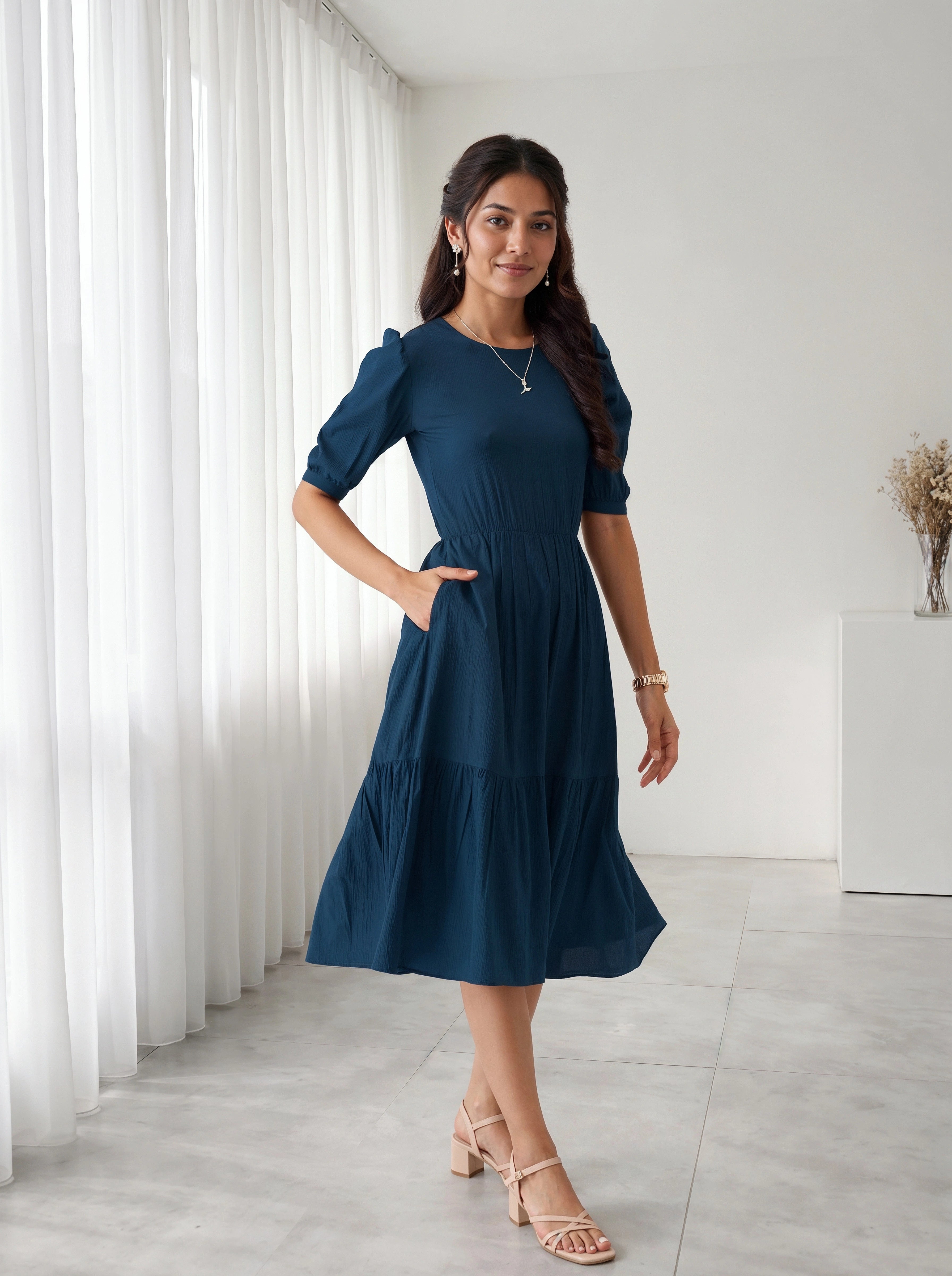 Woman wearing a blue dress standing in a room with white curtains and a white wall.