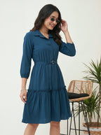 Woman wearing a blue dress with sunglasses, standing indoors with a chair and plant in the background.