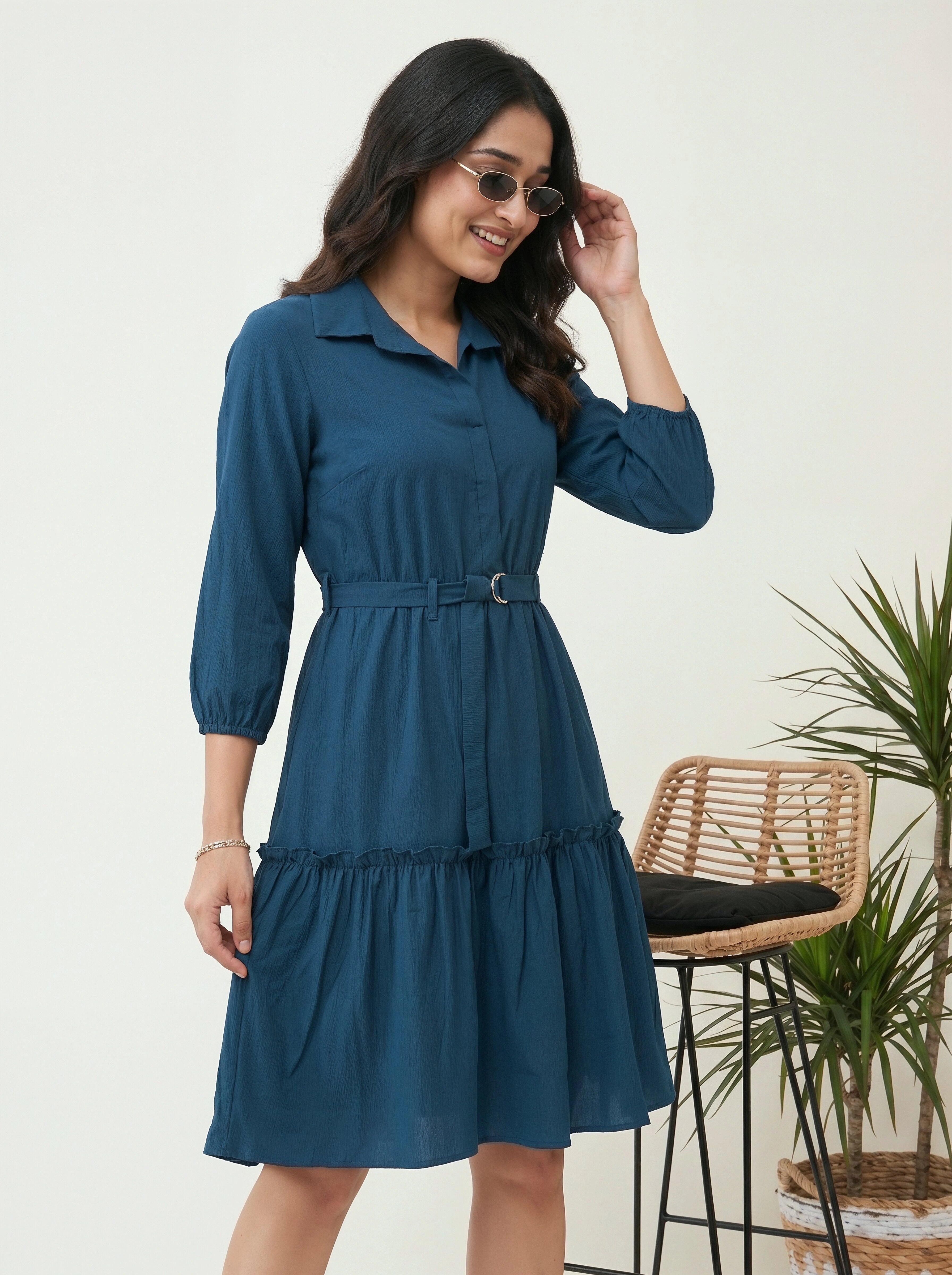 Woman wearing a blue dress with sunglasses, standing indoors with a chair and plant in the background.