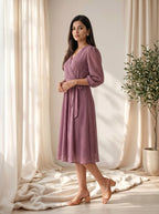 Woman wearing a purple dress standing in a softly lit room with curtains and a plant.