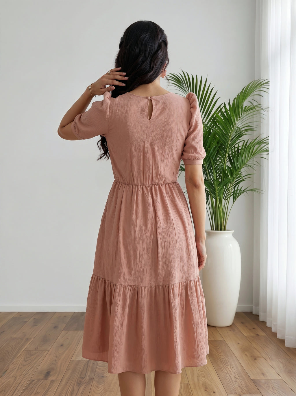 Woman wearing a pink dress standing in a room with a plant and white curtain.