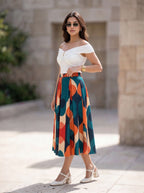 A woman models a stylish off-the-shoulder white top paired with a high-waisted midi skirt featuring a bold geometric pattern in teal, orange, red, and beige. She is wearing white heeled sandals and sunglasses, standing outdoors against a blurred stone wall background.