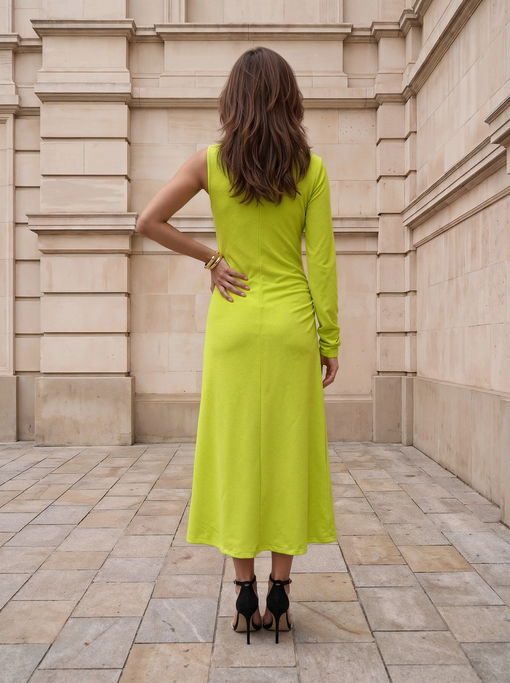 A woman viewed from behind wearing a bright lime green long-sleeve midi dress and black high heels, standing on a stone tiled walkway in front of a beige stone building.