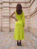 A woman viewed from behind wearing a bright lime green long-sleeve midi dress and black high heels, standing on a stone tiled walkway in front of a beige stone building.