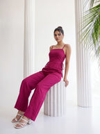 A woman models a magenta two-piece jumpsuit consisting of a spaghetti-strap camisole top and matching wide-leg trousers, sitting on a white fluted pedestal in a bright, modern room with white columns.