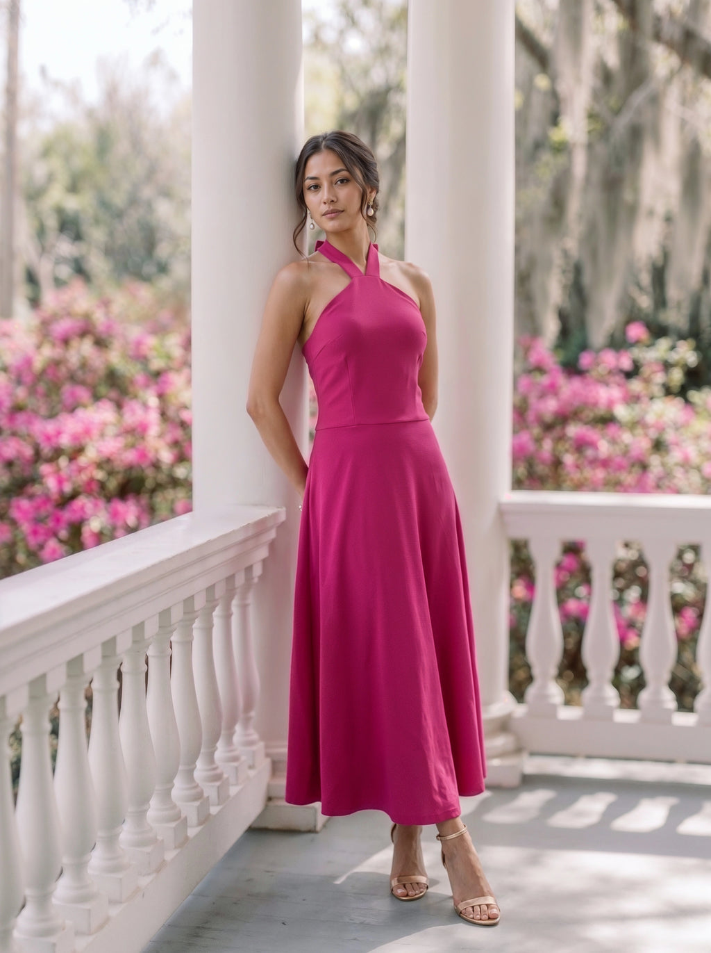A woman stands on a white porch wearing a sleeveless, halter-neck, floor-length magenta dress. The background features white columns, a white railing, and lush green foliage with pink flowers.