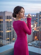 A woman wearing a bright magenta long-sleeve dress stands on a rooftop balcony with a city skyline and blurred lights in the background at dusk.