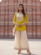 A woman stands in front of the Hawa Mahal palace in Jaipur, wearing a bright yellow kurta with silver mirror work embroidery on the chest, paired with wide-leg cream trousers and carrying a small cream shoulder bag.