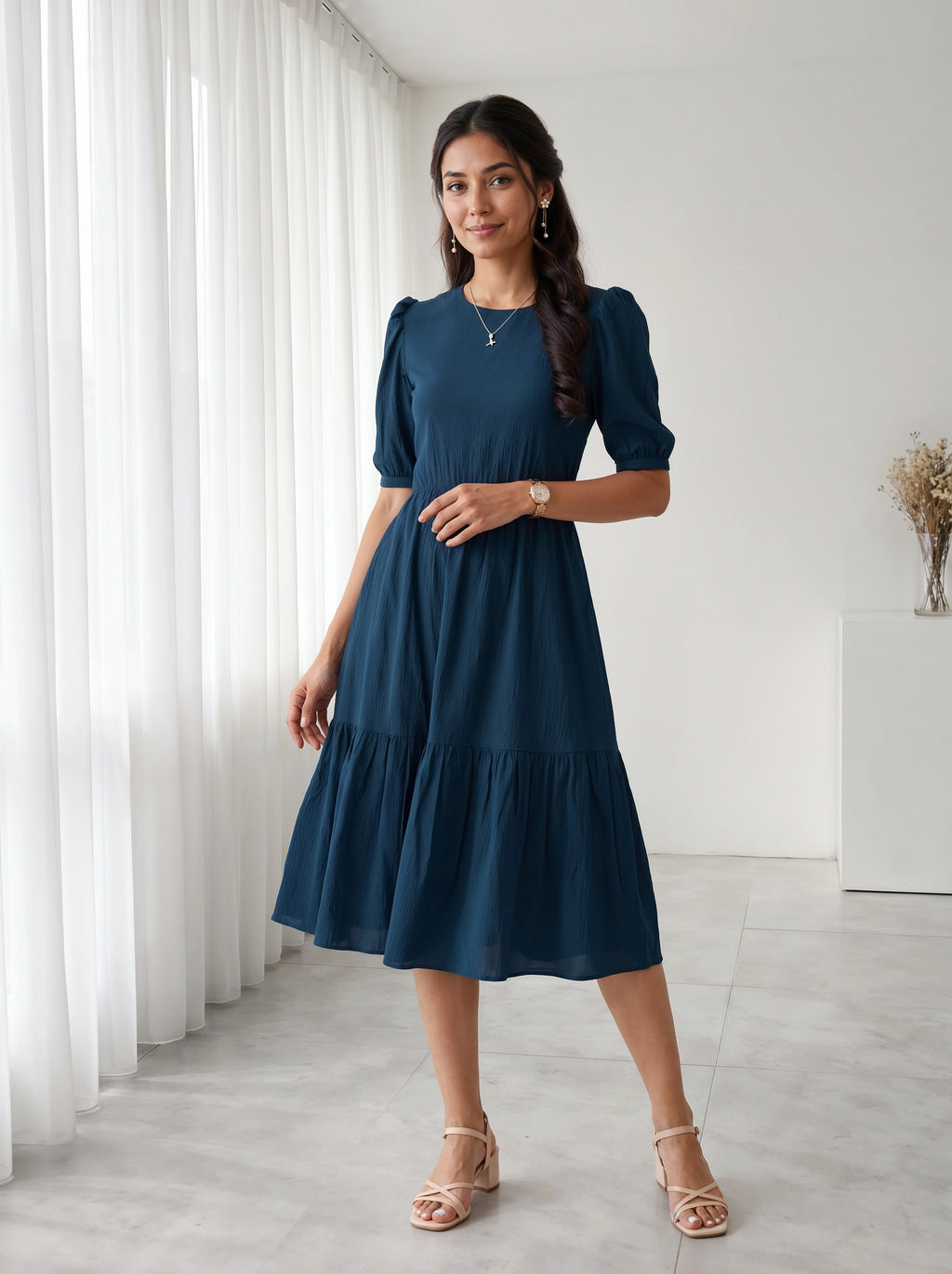 Woman wearing a blue dress standing in a room with white curtains and a white cabinet.