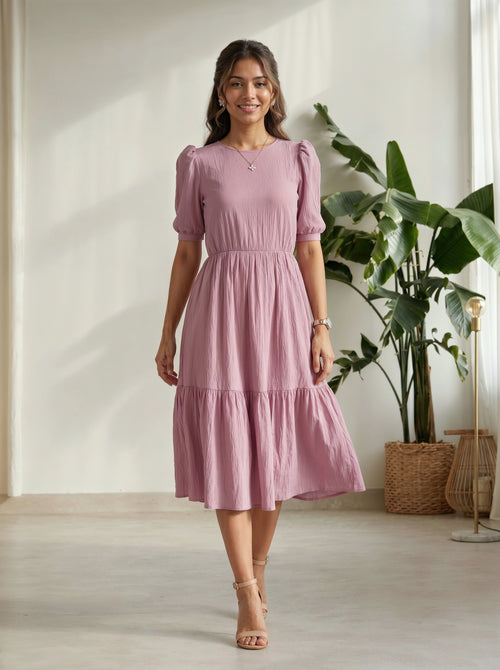 Woman wearing a pink dress standing in a room with plants and a light-colored floor.